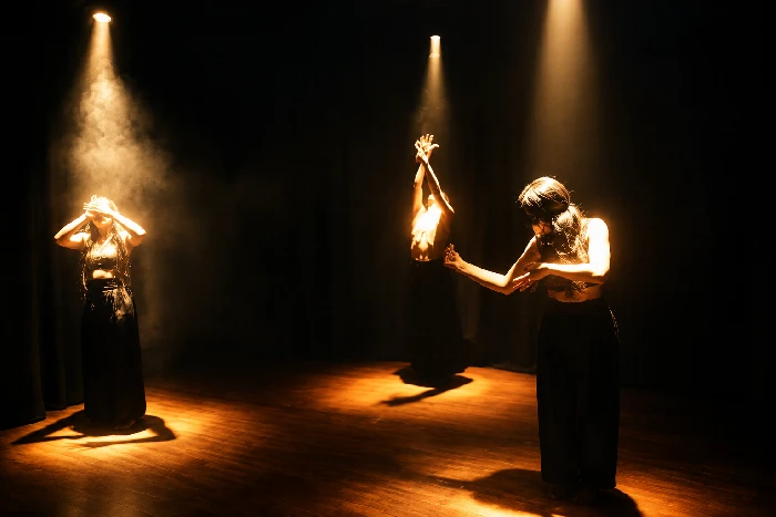 Performance teatral iluminada por haces de luz, con tres intérpretes en escena y una atmósfera de claroscuro.